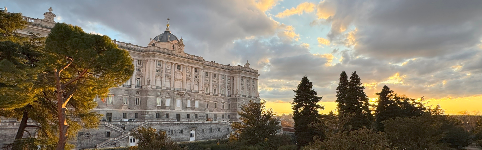 A stone building with a circular tower in front of a yellow and blue sunset peaking through several clouds. 